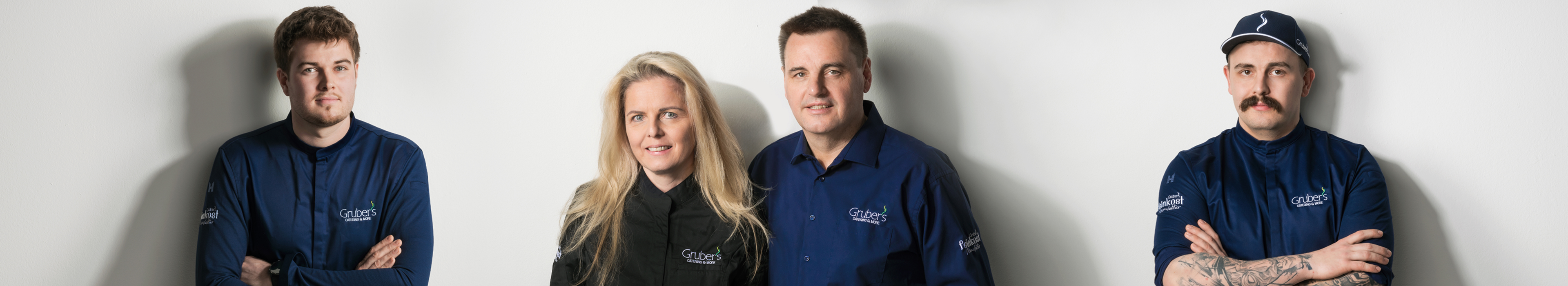 Grubers Feinkost Marufaktur Team Ein Teamfoto der Gruber's Feinkost Manufaktur mit vier Mitarbeitenden in Unternehmenskleidung vor einer weißen Wand.
